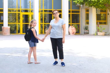 Büyükanne ve öğrenci çocuk okula giderken el ele tutuşuyor. Okul çantalı küçük kız ya da büyükannesiyle okula yürüyen çantalı kız. Yaşlı ebeveyn ve kız, büyükanne ve torun, okula dönüyorlar..