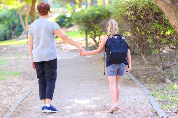 Grandma and pupil kid holding hands going to school.Little girl with school bag or satchel walking to school with grandmother. Old Parent and daughter, grandmother and granddaughter, back view.