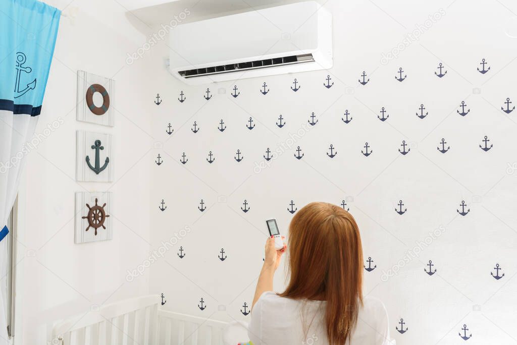 Portrait Of A Happy Woman Holding Remote Control In Front Of Air Conditioner At Home in childrens room.Air conditioner inside the cute baby boys room.With hot weather,is a good solution for family.