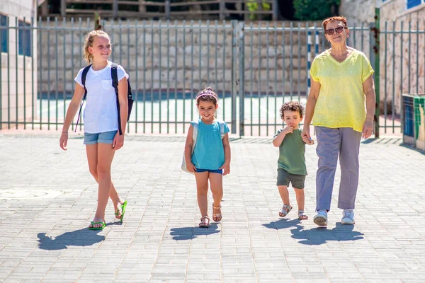 Glückliche Schulkinder, die mit der Großmutter zur Schule gehen.Schüler und Oma mit dem Rucksack gehen in den Kindergarten und in die Schule. — Stockfoto