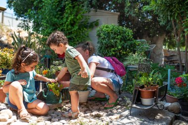 Gerçek hayat. Bahçede çiçek saksılarındaki çiçeklere bakan üç çocuk. Çiçek saksısı yerine bir çaydanlığa sulu bitki saksısı koymak, geri dönüşümlü fikir..