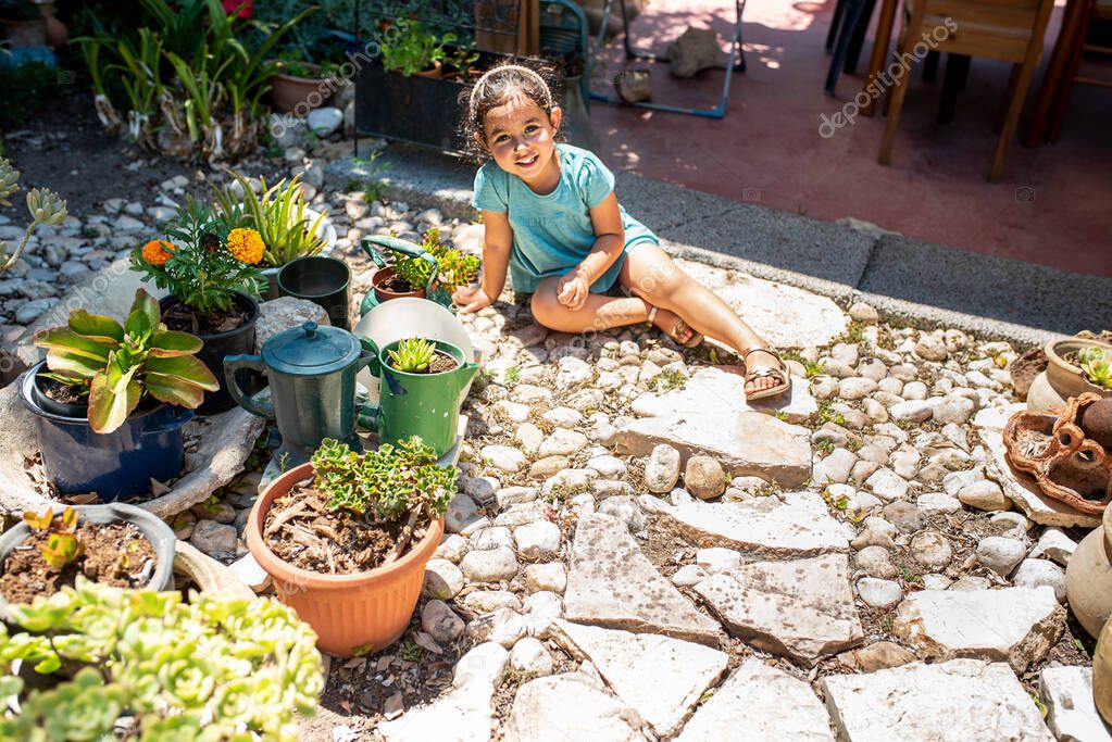 Child playing in the garden. Kids gardening. Little happy girl looking flowers sitting on a stone lawn in the backyard in summer. Toddler kid near potted succulent plants in a teapot, recycled idea.