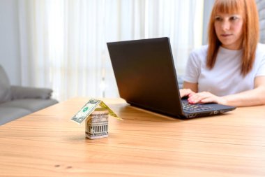 Tahta masadaki dolardan banknota kadar para evi. Orta yaşlı endişeli kadın evdeki oturma odasında dizüstü bilgisayar kullanıyor. Ev satın alma, emlak faaliyetleri, satılık yeni evler arama kavramı..