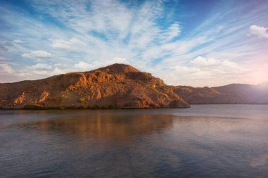 Umman 'lı Jebel Sifah' ın bulutlu mavi gökyüzü ile bir gölün berrak sularında güzel bir dağ manzarası..
