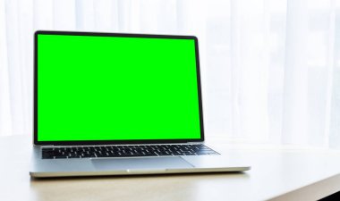Pencerenin önündeki ahşap masa içi yeşil ekranlı dizüstü bilgisayarın maketi ofis odası arka planı, iş dünyası konsepti