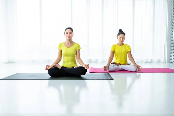 Two young Asian women workout practicing yoga in yellow dress or pose with a trainer and practice meditation wellness lifestyle and health fitness concept in a gym.