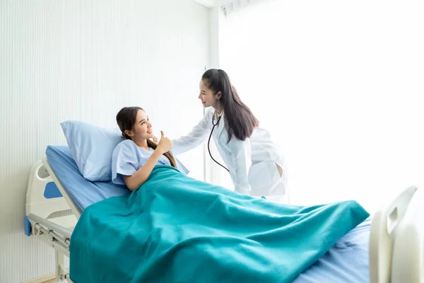 アジアの若い女性患者オンベッドショー親指アップとともにスマイリーフェイス非常に良い症状へアジアの若い女性医師で病院の背景 — ストック写真