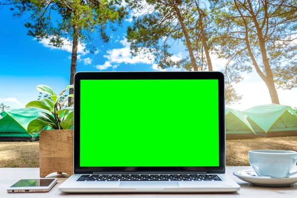 Mockup of laptop computer with empty green screen with coffee cup and smartphone on table in forest summer camp among meadow on Mountain,recreation and Tourist holiday outdoor concept