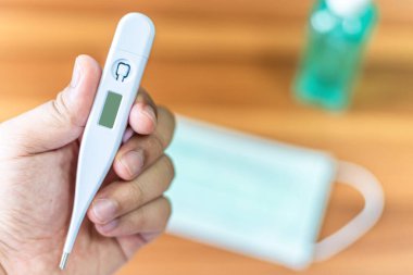 Ev arka plandaki odada, COVID-19 virüsünün yayılmasını önleme kavramında, tahtanın üzerinde tek kullanımlık koruyucu tıbbi maske ve alkol el dezenfektanı ile termometre soyut bulanıklığının el ele tutuşması.