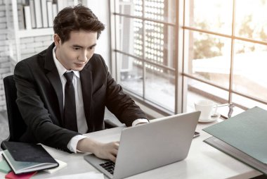 Asyalı genç işadamının başarılı bir iş planını dizüstü bilgisayarda ve tahta masa arkasındaki kalemle gördüğüne sevinen iş dünyası, güveni artırdı.