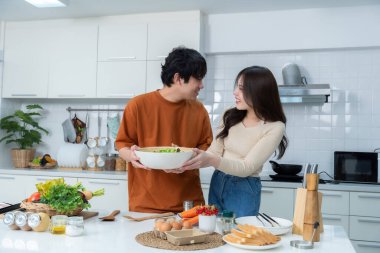 Sevgi dolu genç bir çiftin mutlu portresi. Asyalı neşeli bir şekilde yemek pişirmeyi seviyor. Sebze, et, ekmek pişiriyor.