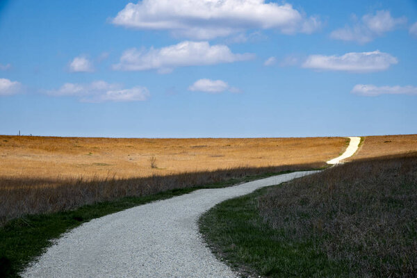 This beautiful Kansas Tallgrass Prairie Preserve photo is patterned with shadows of the clouds overhead and has a winding road disappearing over a hill of early spring golden grass