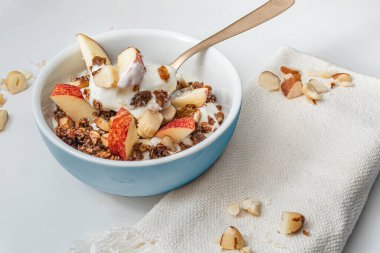 Orta mavi bir kase ev yapımı granola, sade yoğurt, elma ve kestane. Beyaz arka planda havlu peçetesi ve altın kaşıkla.