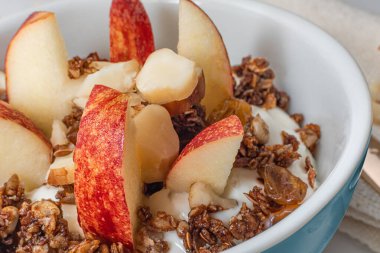 Orta mavi bir kase ev yapımı granola, sade yoğurt, elma ve kestane. Beyaz arka planda havlu peçetesi ve altın kaşıkla.