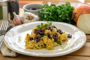 Patates pipetli, karışık yumurtalı ve koyu zeytinli lezzetli bir morina balığı. Tahta bir masada, sarımsak, soğan, maydanoz ve tarifin diğer malzemeleriyle..