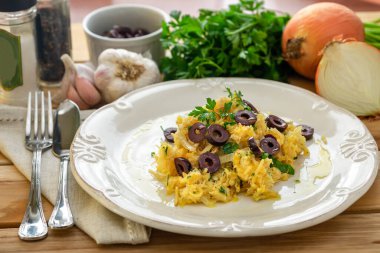 Patates pipetli, karışık yumurtalı ve koyu zeytinli lezzetli bir morina balığı. Tahta bir masada, sarımsak, soğan, maydanoz ve tarifin diğer malzemeleriyle..