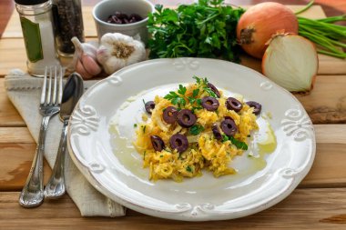 Patates pipetli, karışık yumurtalı ve koyu zeytinli lezzetli bir morina balığı. Tahta bir masada, sarımsak, soğan, maydanoz ve tarifin diğer malzemeleriyle..