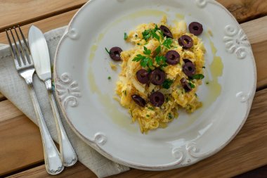 Patates pipetli, karışık yumurtalı ve koyu zeytinli lezzetli bir morina balığı. Tahta bir masada, sarımsak, soğan, maydanoz ve tarifin diğer malzemeleriyle..