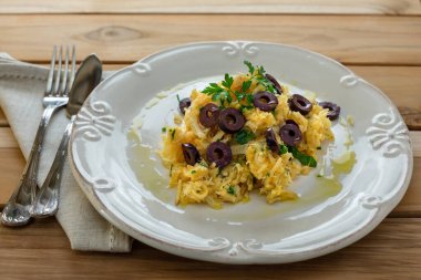 Patates pipetli, karışık yumurtalı ve koyu zeytinli lezzetli bir morina balığı. Tahta bir masada, sarımsak, soğan, maydanoz ve tarifin diğer malzemeleriyle..