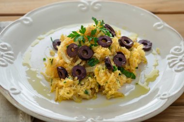 Patates pipetli, karışık yumurtalı ve koyu zeytinli lezzetli bir morina balığı. Tahta bir masada, sarımsak, soğan, maydanoz ve tarifin diğer malzemeleriyle..