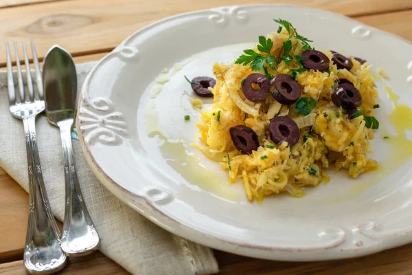 Patates pipetli, karışık yumurtalı ve koyu zeytinli lezzetli bir morina balığı. Tahta bir masada, sarımsak, soğan, maydanoz ve tarifin diğer malzemeleriyle..