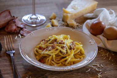 Lezzetli bir İtalyan Carbonara makarnası, ahşap bir masada, rendelenmiş parmesan peyniri, dilimlenmiş pastırma, yanında iki yumurta ve bir bardak şarap ile servis edilir..