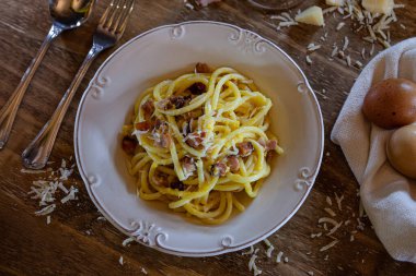 Lezzetli bir İtalyan Carbonara makarnası, ahşap bir masada, rendelenmiş parmesan peyniri, dilimlenmiş pastırma, yanında iki yumurta ve bir bardak şarap ile servis edilir..