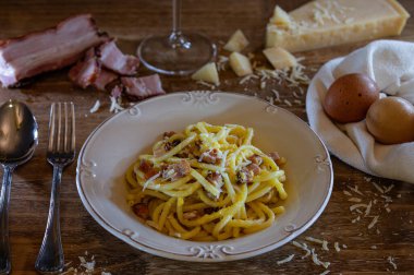 Lezzetli bir İtalyan Carbonara makarnası, ahşap bir masada, rendelenmiş parmesan peyniri, dilimlenmiş pastırma, yanında iki yumurta ve bir bardak şarap ile servis edilir..
