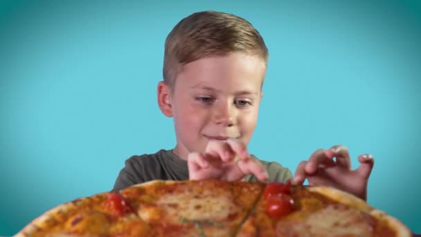 Lindo niño comiendo pizza con placer en un fondo azul colorido — Vídeo de stock