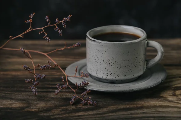 Koyu arkaplanda, ahşap bir masada bir fincan espresso kahvesi. Yakınlarda kurumuş bir çiçek fidanı.