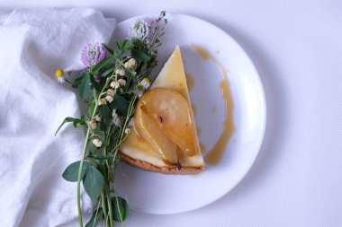 New York cheesecake Karamelize armut ile hizmet vermiştir. Beyaz plaka üzerinde yaban çiçekleri ve beyaz keten bez ile süslenmiş cheesecake