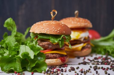 İki büyük klasik Amerikan cheeseburger. Farklı biber ve marul çeşitleri ile dekore edilmiş Cheeseburgerler. Koyu ahşap arka plan 