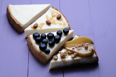 Mor ahşap arka planda farklı hizmet seçenekleri ile New York cheesecake dilimleri. Karamel ve badem. Armut, karamel ve badem. Yaban mersini. Klasik