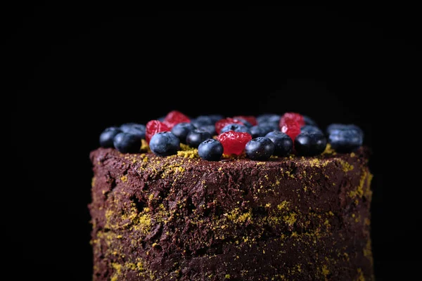 Chocolate cake with blueberries and dried cherries decorated with ground pistachios and berries on a dark background.