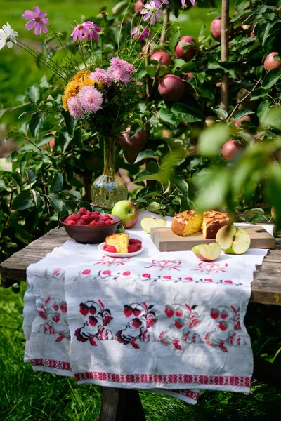 A piece cut from an apple pie lies on a plate. Rustic still life: sunny bright day, apple pie under an apple tree on an old wooden table, fruits and berries, flowers.
