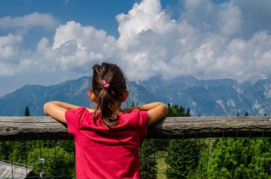Bir çitin yaslanmış genç kız, bulutlar ve mavi gökyüzü ile dağlara bakıyor