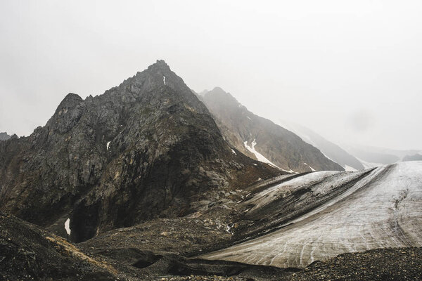 Высокая перимидальная гора рядом с тающим ледником. Скалистые склоны покрыты снегом. Объемная фотография Алтая. Острые пики скрыты непроницаемым туманом. Начало горных рек
.