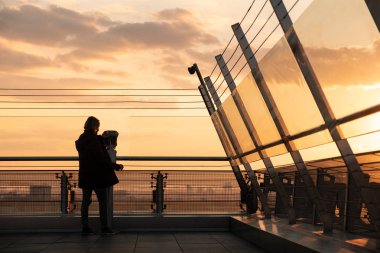 Dürbünle gün batımını izleyen bir adamın silueti. Bir gökdelenin çatısında gözlem güvertesi. Güneş bulutların arasından parlıyor. Kızıl Gök. Dürbünü eliyle tutuyor..
