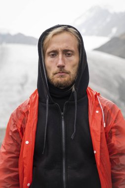 Buzulun arka planındaki kasvetli adam. Kırmızı yağmurluk, siyah kalın kıyafetler. Gergin yüzlü bir turistin portresi. Sarışın, mavi gözlü, sakallı. Altai Dağı. Soğuk kar.