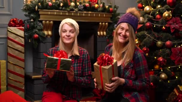 dvě legrační mladé dívky v červených košilích a legrační klobouky sedět v blízkosti Nového roku nebo vánoční stromeček a vyměnit dárky