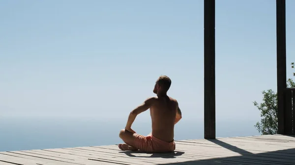 Slow pan footage of man doing yoga in a special wooden sports house. Handsome muscular bearded man in shorts raises his arms up. Businessman on vacation