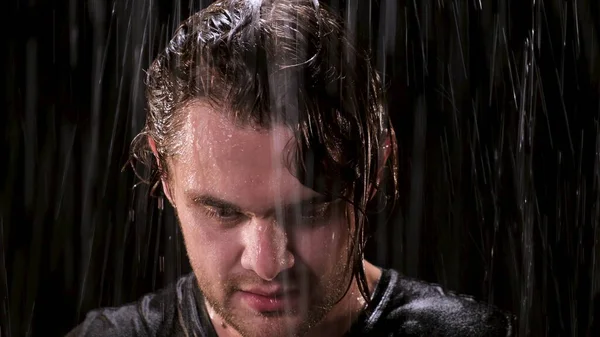 Sad guy with long hair stands in the rain at night and looks up at the camera. Close up eyes of man are full of bitterness in bad weather.