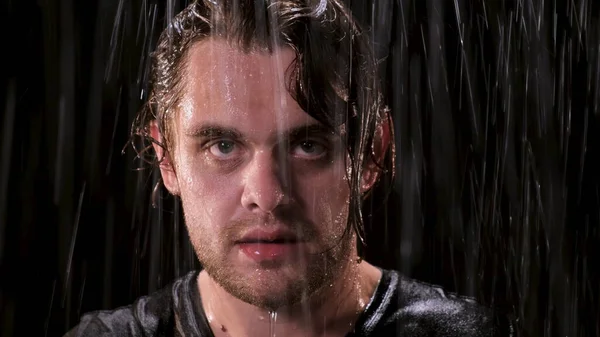 Sad guy with long hair stands in the rain at night and looks up at the camera. Close up eyes of man are full of bitterness in bad weather.