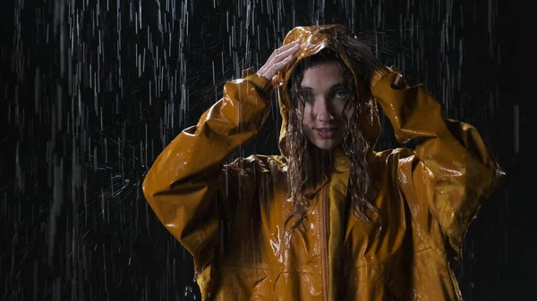 Attractive young woman in yellow raincoat stands on the street at night in the rain. Cute girl straightens her hood so as not to get wet and looks at the camera.