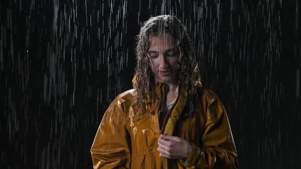 Satisfied woman in yellow raincoat puts on hood raincoat. It is pouring rain, the woman is all wet, she has beautiful wavy wet hair. Lady stands in the street at night.
