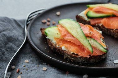 Krem peynir, somon ve salatalık ile açık sandviç yakın çekim gri arka plan üzerinde koyu ahşap plaka kaplı koyu gri peçete ve plaka yakınında çatal ile pembe deniz tuzu