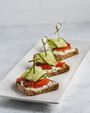 Beyaz arka planda beyaz dikdörtgen plaka üzerinde krem peynir, somon ve salatalık ile üç bruschettas