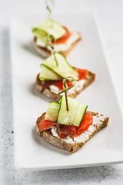 Beyaz arka planda beyaz dikdörtgen plaka üzerinde krem peynir, somon ve salatalık ile üç bruschettas