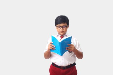 Endonezya ilkokulu üniformalı Asyalı bir öğrencinin portresi ayakta duruyor ve ciddi ciddi kitap okuyor. Gri arkaplanda izole