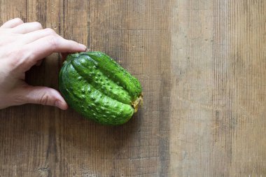 İnsan eli, çirkin üçlü yeşil organik salatalığı tutuyor. Kahverengi ahşap arka planda ve fotokopi alanı olan alışılmadık şekilli sebzeler. Gıda atığını azaltmak için kusurlu ürün satın almak. Çevre trendi. Yatay.